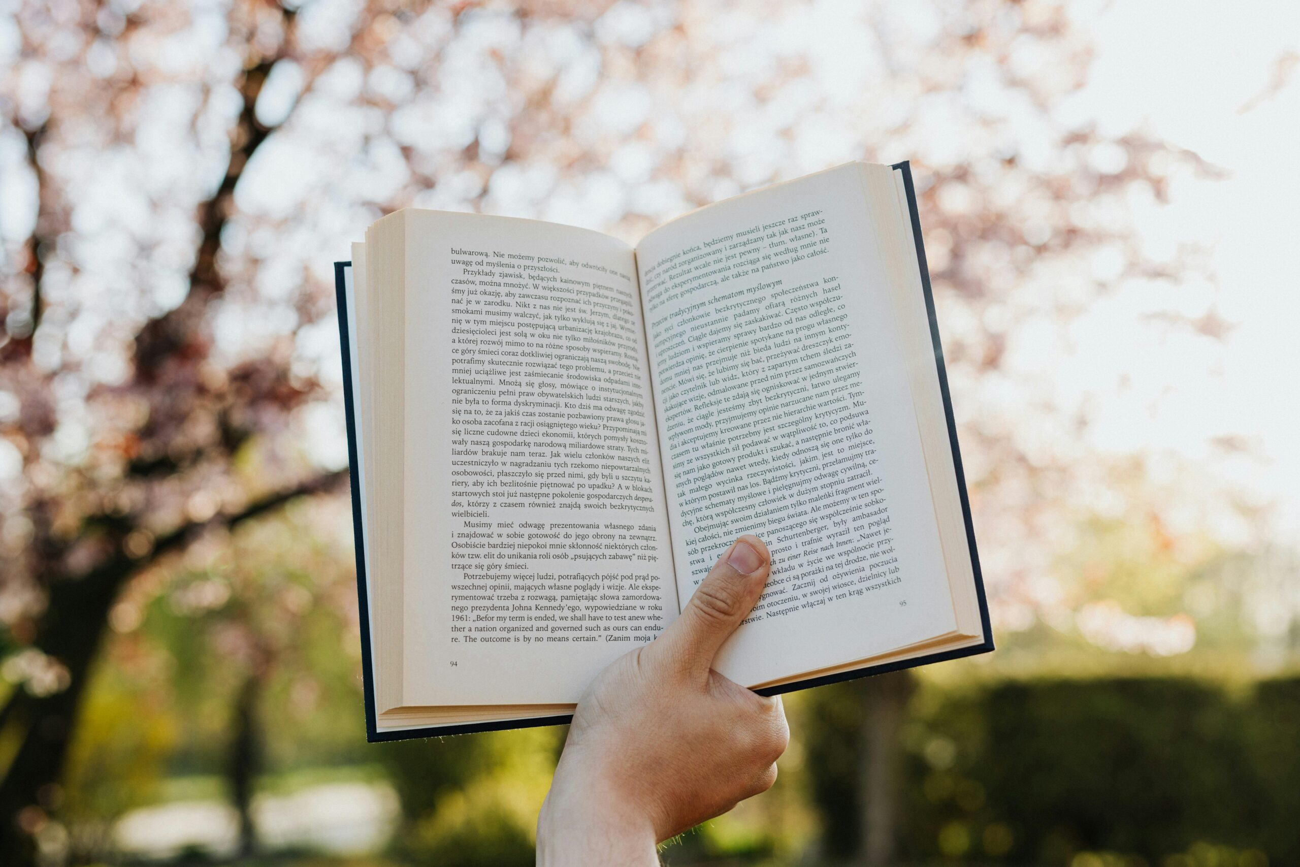 A hand holds an open book with blurred spring blossoms in the background.
