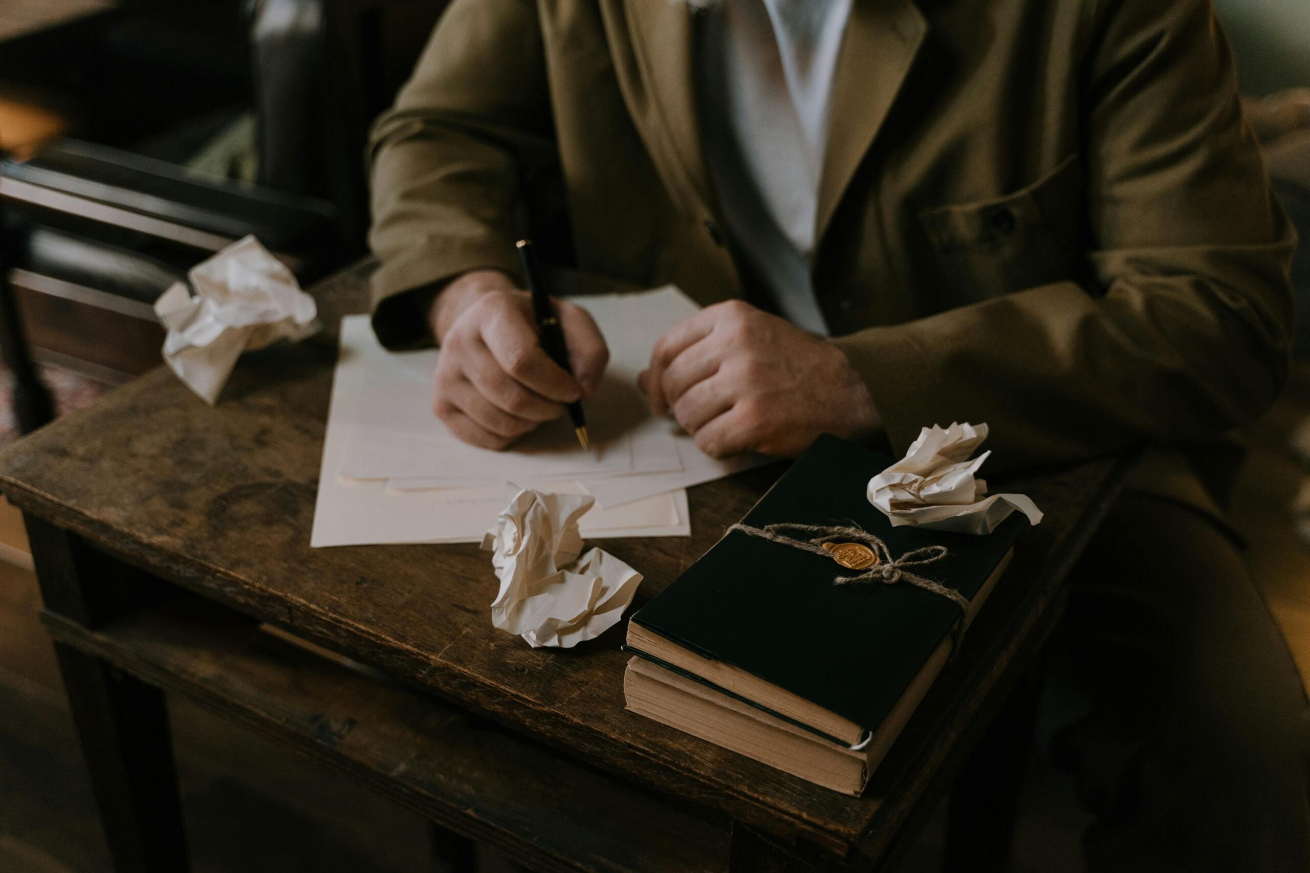 A person writing at a wooden desk with crumpled papers and a book.