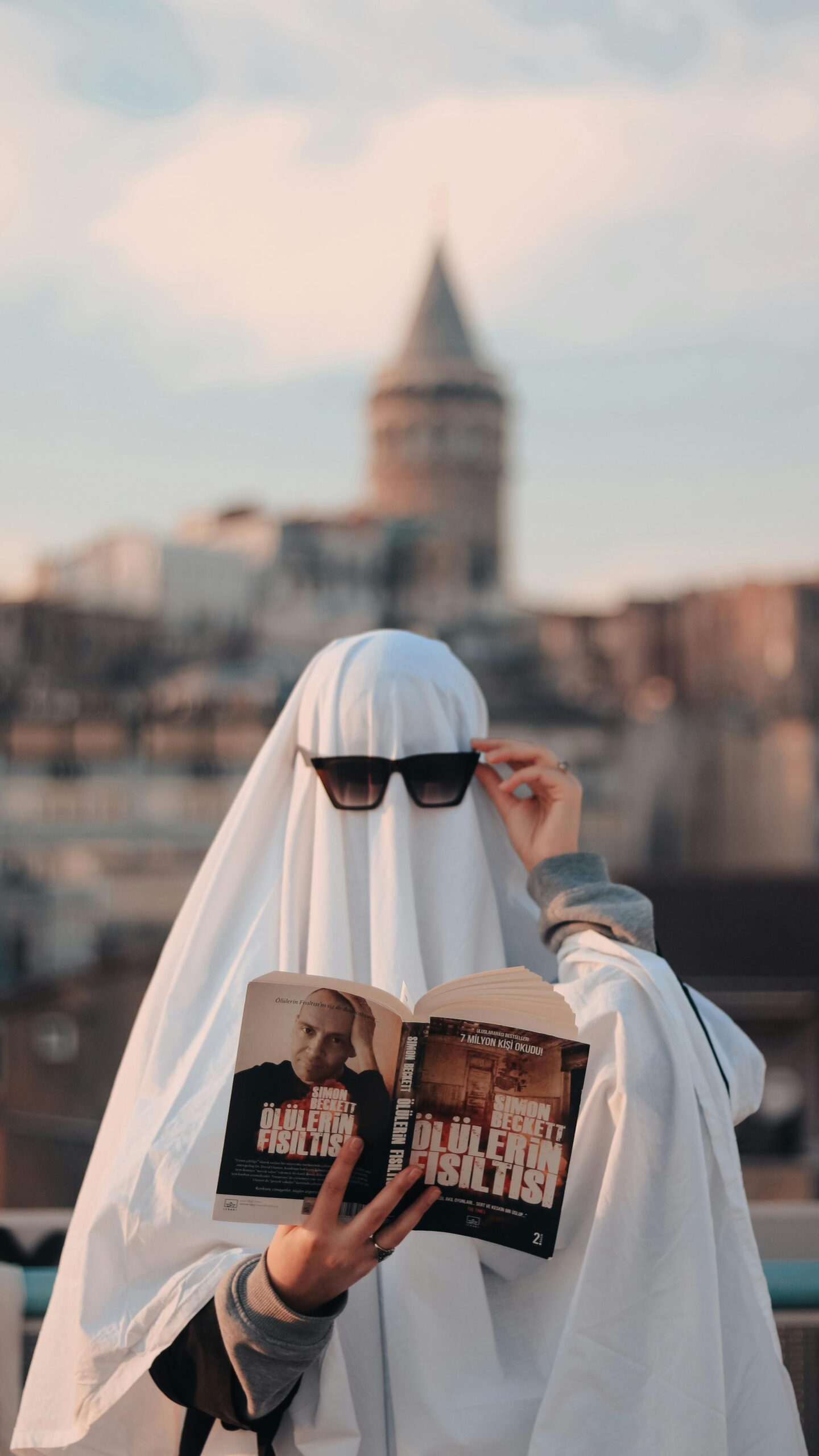 A person wearing a ghost costume reads a book outdoors near a historic tower.